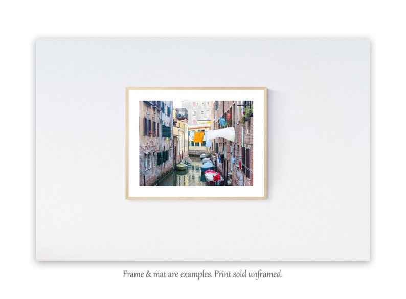 May include: A photograph of a narrow canal in Venice, Italy. The canal is lined with colorful buildings and there are several boats docked along the sides. Laundry is hanging from lines strung between the buildings.