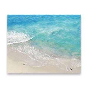 May include: A high-angle view of a beach with turquoise water and white waves. The sand is a light beige color. The ocean transitions from a deep blue to a lighter turquoise as it approaches the shore. The image is a photograph.