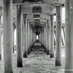 Pode incluir: Uma fotografia a preto e branco de um cais com muitos pilares de bet&atilde;o que se estendem para a &aacute;gua. Os pilares criam um efeito de t&uacute;nel, que leva a uma luz brilhante no final.
