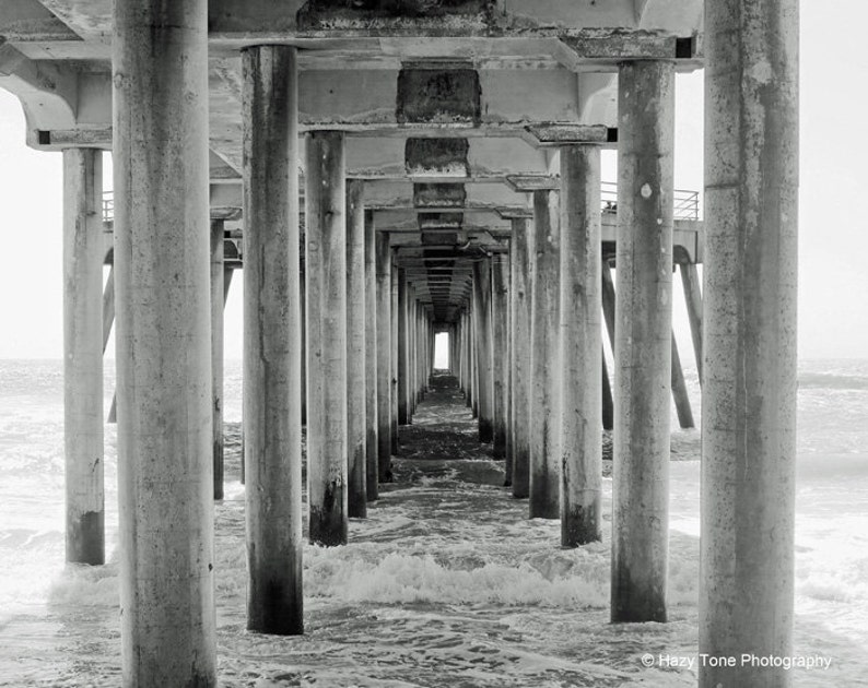 Pode incluir: Uma fotografia a preto e branco de um cais com muitos pilares de bet&atilde;o que se estendem para a &aacute;gua. Os pilares criam um efeito de t&uacute;nel, que leva a uma luz brilhante no final.