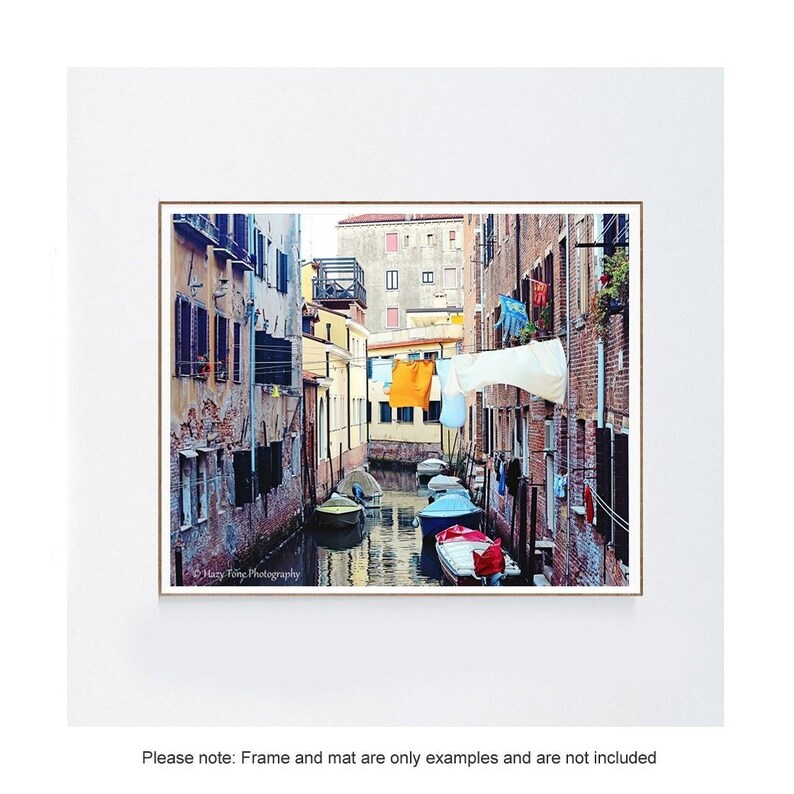May include: A narrow canal in Venice, Italy, with colorful buildings lining the sides. Laundry is hanging on a clothesline strung across the canal. Several small boats are docked along the canal.