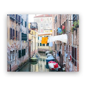 May include: A narrow canal in Venice, Italy, with colorful buildings lining the sides.  Laundry is hanging on lines strung across the canal.  Several small boats are docked in the canal.