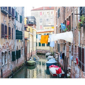 May include: A narrow canal in Venice, Italy, with colorful laundry hanging from lines strung between buildings.  The buildings are old and have a rustic, weathered look.  Several small boats are docked in the canal.