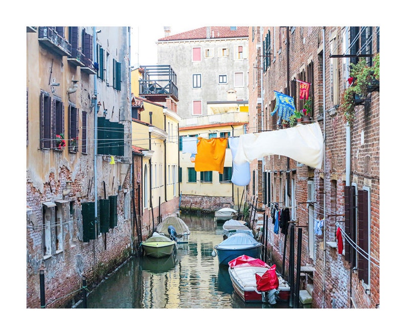 May include: A narrow canal in Venice, Italy, with colorful laundry hanging from lines strung between buildings.  The buildings are old and have a rustic, weathered look.  Several small boats are docked in the canal.