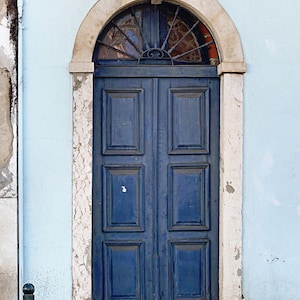 May include: A blue wooden double door with a decorative iron grate above the door. The door is set in a white stone archway against a light blue wall.