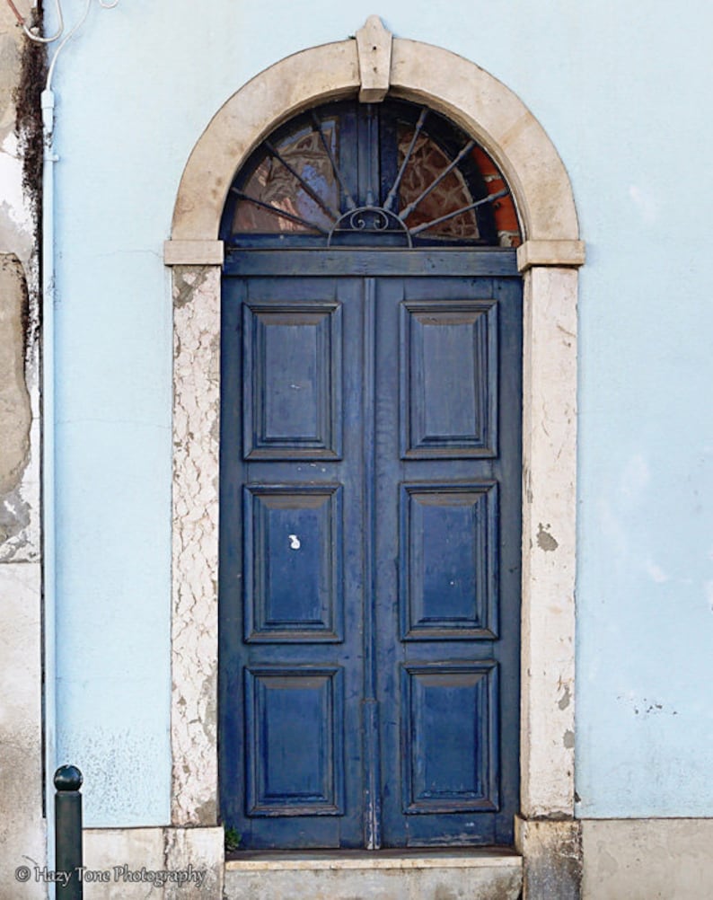 May include: A blue wooden double door with a decorative iron grate above the door. The door is set in a white stone archway against a light blue wall.