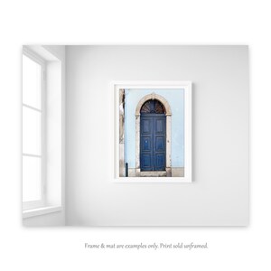 May include: A framed photograph of a blue door with an arched top, set against a light blue wall. The door is made of wood and has a simple design. The photograph is in a white frame.