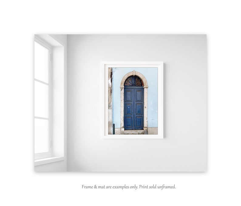 May include: A framed photograph of a blue door with an arched top, set against a light blue wall. The door is made of wood and has a simple design. The photograph is in a white frame.