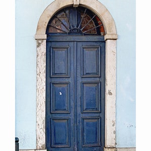May include: A blue wooden double door with a decorative iron grate above it. The door is set in a stone archway and is framed by white stone trim. The door is surrounded by a light blue wall.