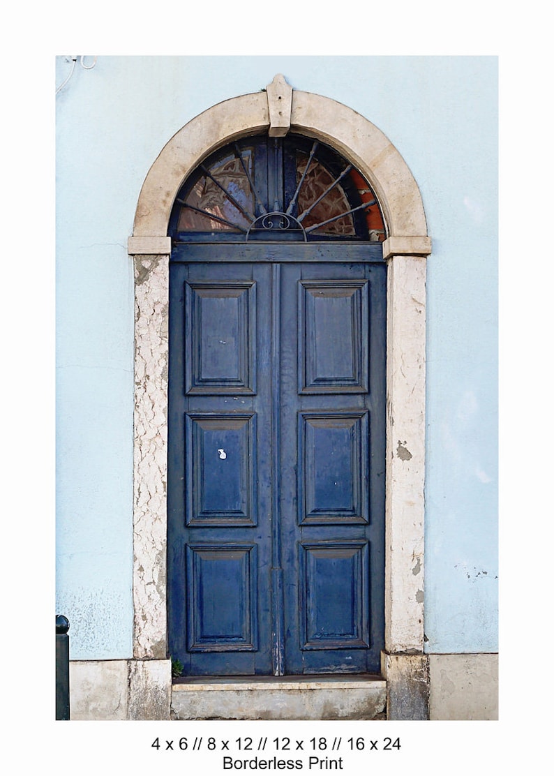 May include: A blue wooden double door with a decorative iron grate above it. The door is set in a stone archway and is framed by white stone trim. The door is surrounded by a light blue wall.