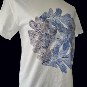 May include: White t-shirt featuring a large leaf design in shades of blue and gray. The leaf print is centered on the front of the shirt. The shirt has short sleeves and a crew neck.