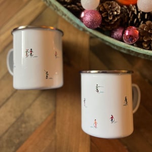 May include: Two white enamel mugs with silver rims and handles. Each mug features a whimsical illustration of skiers in various colourful outfits. The mugs are set on a wooden surface, with a decorative tray of pinecones and baubles in the background.