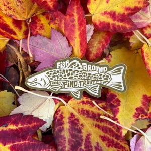 Op de afbeelding: Een sticker met een visillustratie en de tekst "Fish Around and Find Trout" ligt op kleurrijke herfstbladeren. De bladeren vertonen tinten rood, geel en paars.