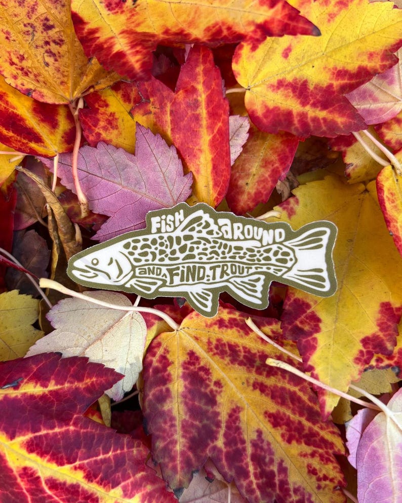 Op de afbeelding: Een sticker met een visillustratie en de tekst "Fish Around and Find Trout" ligt op kleurrijke herfstbladeren. De bladeren vertonen tinten rood, geel en paars.
