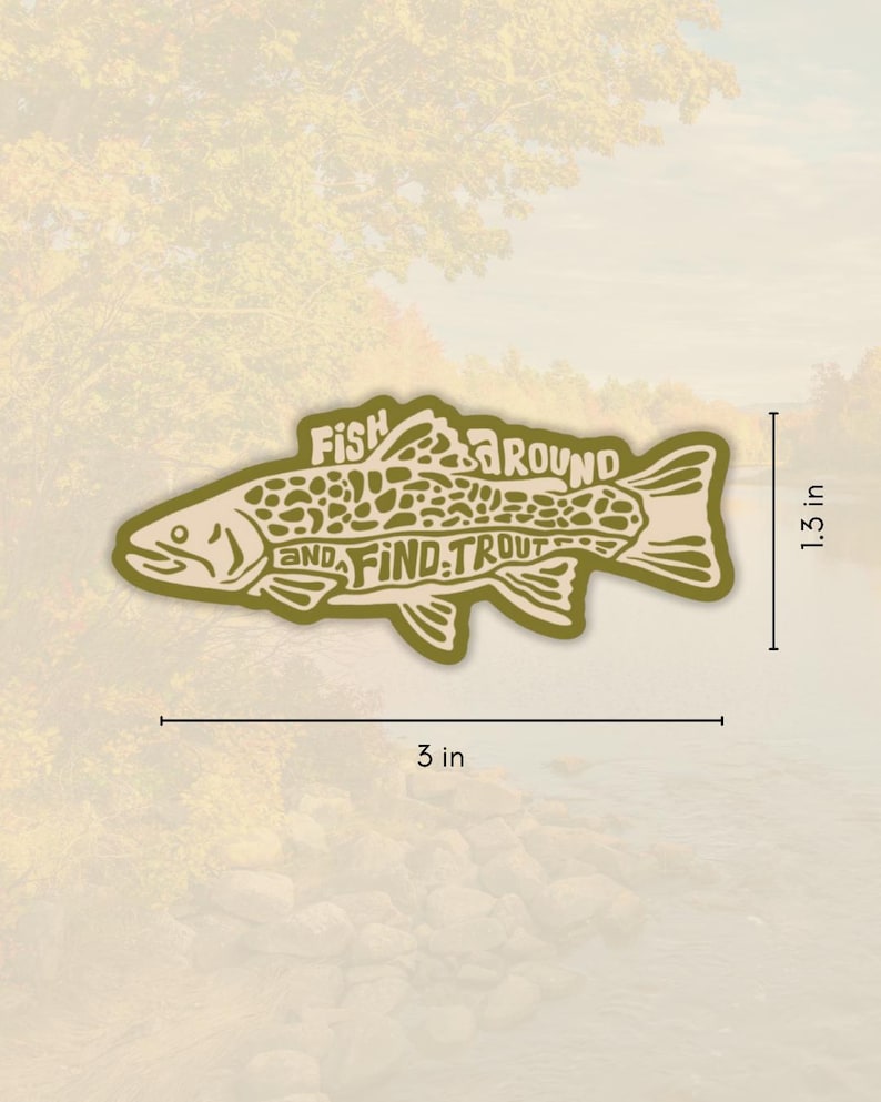 Op de afbeelding: Een gestanste sticker in de vorm van een vis, met de woorden "Fish Around and Find Trout" in een gestileerd lettertype. De sticker is ongeveer 7,6 cm lang en 3,3 cm hoog, met een lichtgroen en beige kleurenschema. De achtergrond is een vage afbeelding van een rivier en bomen.