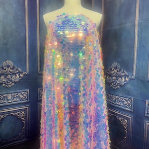 May include: A sleeveless garment draped over a mannequin, entirely covered in iridescent, circular sequins that reflect light in shades of pink, blue, and gold. The sequins are arranged in a dense, overlapping pattern, creating a shimmering effect.
