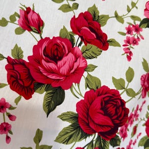 May include: Close-up of a white fabric with a repeating floral pattern. Large, vibrant red roses with pink centers and green leaves are scattered across the surface. Smaller pink flowers and green vines fill the spaces between the roses.
