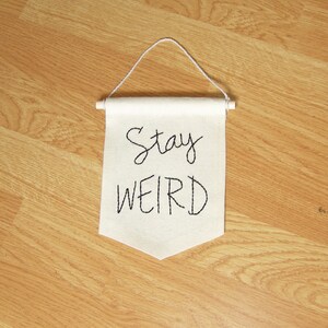 Może przedstawiać: Kremowy baner z wyhaftowanymi czarnymi literami "Stay WEIRD". Baner zawieszony jest na białym sznurku i drewnianym kołku. Baner jest wyświetlany na drewnianej podłodze.