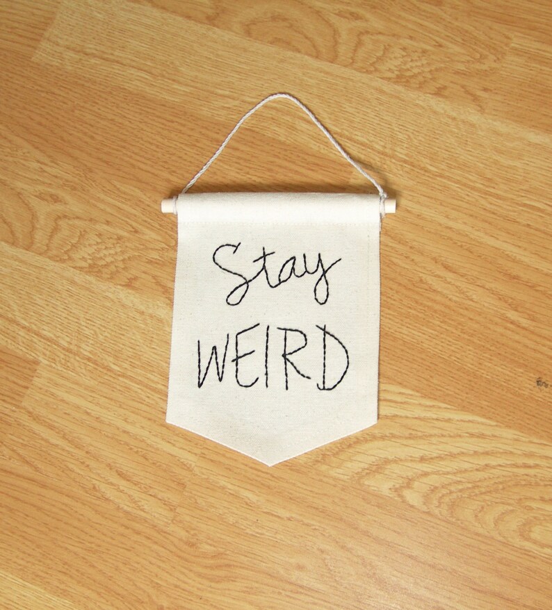Może przedstawiać: Kremowy baner z wyhaftowanymi czarnymi literami "Stay WEIRD". Baner zawieszony jest na białym sznurku i drewnianym kołku. Baner jest wyświetlany na drewnianej podłodze.