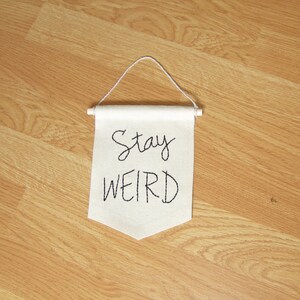 Może przedstawiać: Mały, kremowy baner z napisem "Stay WEIRD" wyhaftowanym czarnym pismem. Baner zawieszony jest na drewnianym kołku z białym sznurkiem do zawieszenia. Baner jest wyświetlany na jasnobrązowej drewnianej powierzchni.