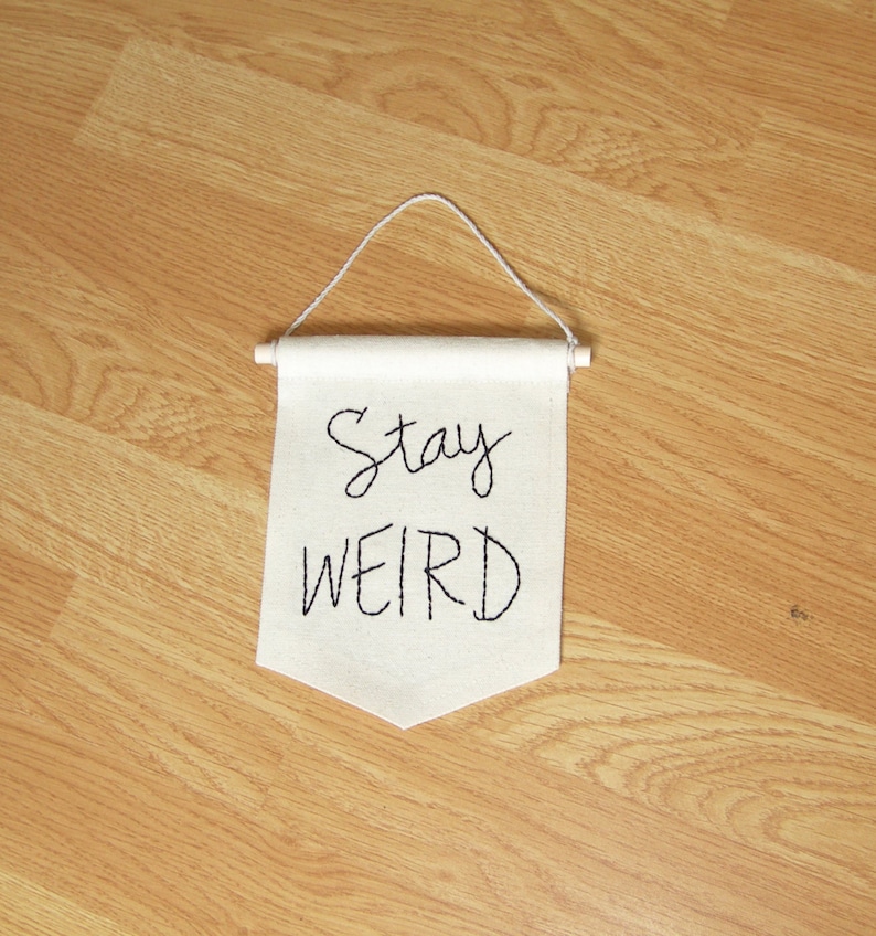 Może przedstawiać: Mały, kremowy baner z napisem "Stay WEIRD" wyhaftowanym czarnym pismem. Baner zawieszony jest na drewnianym kołku z białym sznurkiem do zawieszenia. Baner jest wyświetlany na jasnobrązowej drewnianej powierzchni.
