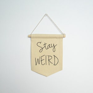 Może przedstawiać: Beżowy baner z tkaniny z wyhaftowanymi czarnymi literami "Stay WEIRD". Baner jest prostokątny ze spiczastym dnem i zawieszony na drewnianym kołku i sznurku.
