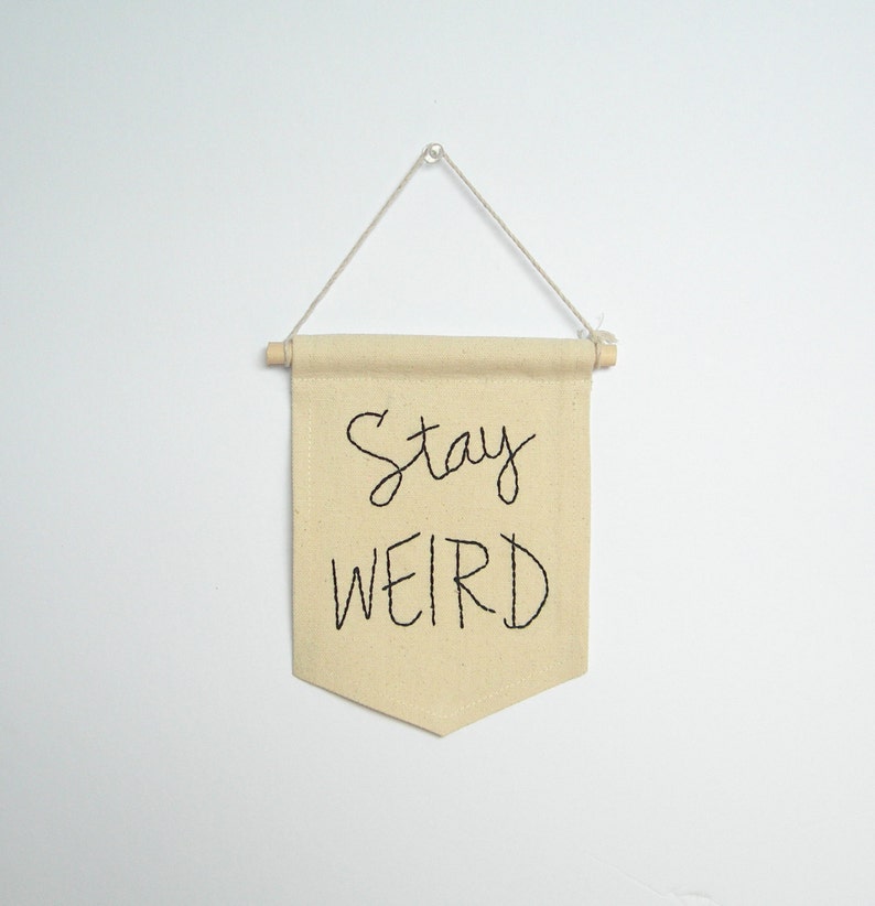 Może przedstawiać: Beżowy baner z tkaniny z wyhaftowanymi czarnymi literami "Stay WEIRD". Baner jest prostokątny ze spiczastym dnem i zawieszony na drewnianym kołku i sznurku.