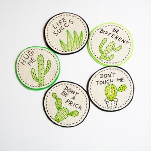 May include: Embroidered circular patches featuring cactus designs and phrases such as "Hug Me" and "Be Different." The patches have a cream background, green and black stitching, and are arranged in a circular pattern.