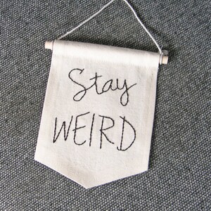 Może przedstawiać: Kremowy baner z wyhaftowanymi czarną nicią słowami "Stay WEIRD". Baner jest prostokątny z ostrym dnem i zawieszony na białym sznurku i drewnianym kołku.