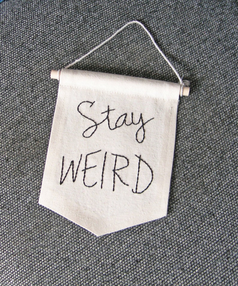 Może przedstawiać: Kremowy baner z wyhaftowanymi czarną nicią słowami "Stay WEIRD". Baner jest prostokątny z ostrym dnem i zawieszony na białym sznurku i drewnianym kołku.