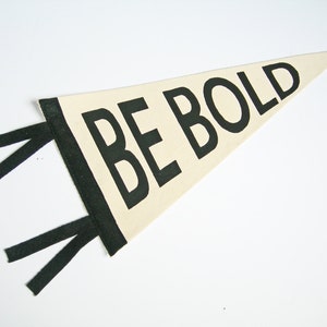 Può includere: Gagliardetto color crema con le parole "BE BOLD" in grassetto nero. La bandiera ha un bordo nero e strisce di tessuto nero che si estendono dal lato sinistro. Il gagliardetto è di forma triangolare.