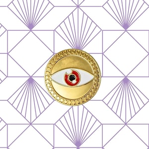 Könnte beinhalten: Goldfarbener Pin mit einem weißen Kreis und einem roten Auge in der Mitte. Der Pin befindet sich auf einem weißen Hintergrund mit einem geometrischen Muster.