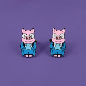 May include: A pair of cufflinks featuring a cartoon pig wearing a blue suit and a white bow tie. The pig has pink skin and a white snout.