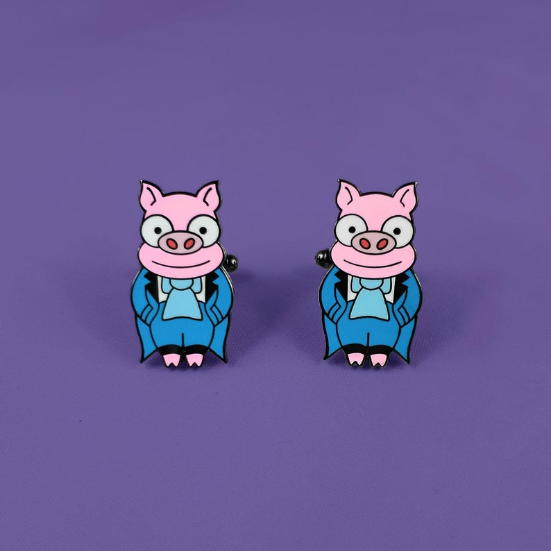 May include: A pair of cufflinks featuring a cartoon pig wearing a blue suit and a white bow tie. The pig has pink skin and a white snout.