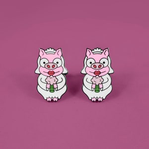 May include: Two pink pig bride enamel pins with white dresses and pink flowers. The pigs have black eyes, black eyelashes, and red lips.