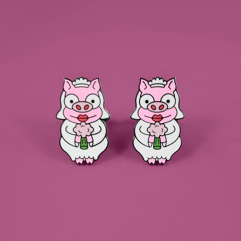 May include: Two pink pig bride enamel pins with white dresses and pink flowers. The pigs have black eyes, black eyelashes, and red lips.