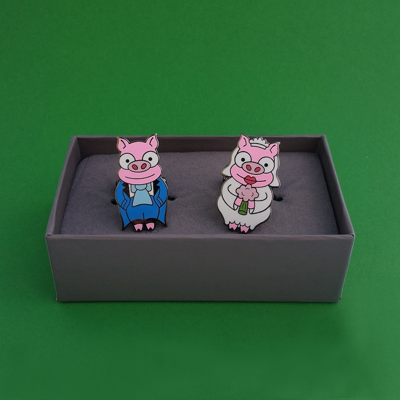 May include: Two pig pins, one wearing a blue suit and the other wearing a white dress and holding a pink flower. The pins are displayed on a gray box.