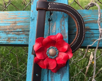 May include: A brown leather horse halter with a red leather flower accent. The flower has a turquoise and copper center with rhinestones.