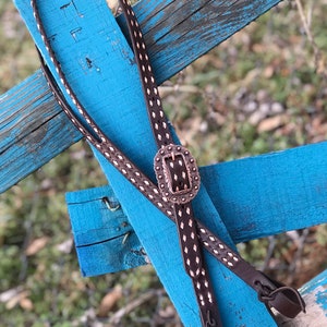 Puede incluir: Un cabezada de cuero marrón para caballo con un patrón de puntadas en forma de diamante y una hebilla plateada y de cobre. La cabezada está colgada de una valla de madera azul.