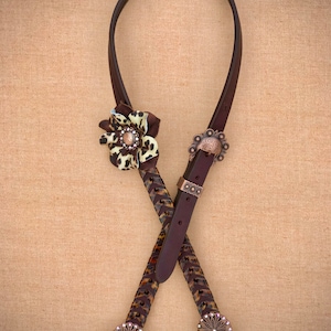 May include: A brown leather horse halter with a braided design and a leopard print accent. The halter has two decorative metal buckles with rhinestones.