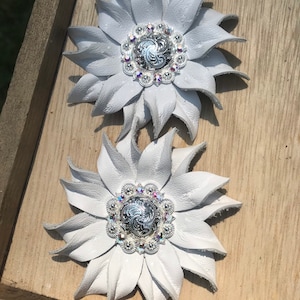 Op de afbeelding: Twee witte lederen bloemenvormen met zilveren en kristallen versieringen. Elke bloem heeft een zilveren midden met een swirl-ontwerp en is omgeven door een cirkel van heldere kristallen.