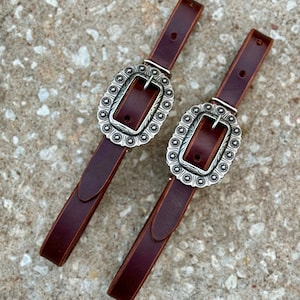 May include: A pair of brown leather spur straps, each featuring an ornate silver-coloured buckle. The buckles are oval and decorated with a floral pattern. The straps are a deep brown, reflecting a classic Western style.