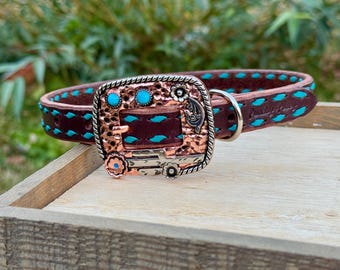 May include: A brown leather dog collar with turquoise stitching and accents. The collar features a decorative copper buckle with turquoise stones and floral details. The buckle is silver-toned with a decorative design. The collar is displayed on a wooden surface.