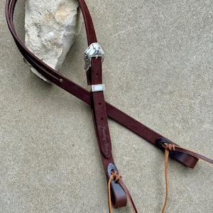 Puede incluir: Una rienda de caballo de cuero marrón oscuro con una hebilla plateada con un diseño de rayo. La rienda tiene un lazo en el extremo y las palabras "Punchy Pony" están estampadas en el cuero.