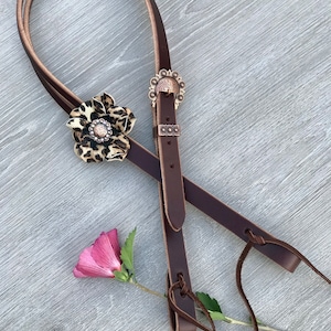 May include: A brown leather halter with a leopard print flower and a decorative concho. The halter has two leather ties at the bottom.