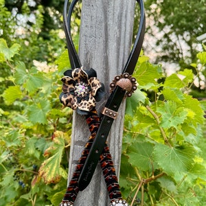 May include: A black leather strap with a leopard print design and a flower accent. The strap has two decorative metal buckles with a floral design.