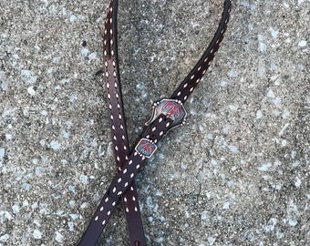 May include: A brown leather strap with white stitching and a silver concho with red and white accents. The strap is designed for a saddle or other equestrian equipment.