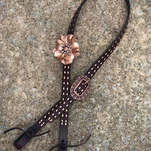Puede incluir: Un par de riendas de cuero marrón con una flor y una hebilla de color cobre. Las riendas están cosidas con un patrón de diamante y tienen un acabado de color cobre.