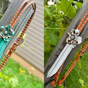 May include: Two brown leather straps with white polka dots and a flower charm. One strap has a turquoise tassel and the other has a white tassel. The flower charm has a leopard print and a rhinestone center.