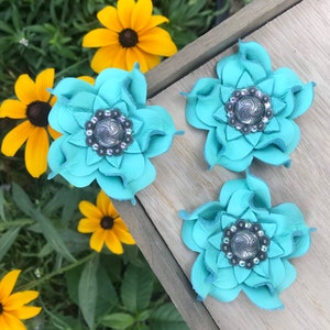 May include: Three turquoise leather flower hair clips with silver and crystal center embellishments. The flower petals are layered, creating a dimensional effect. The clips are arranged on a wooden surface, with yellow flowers and green foliage in the background.